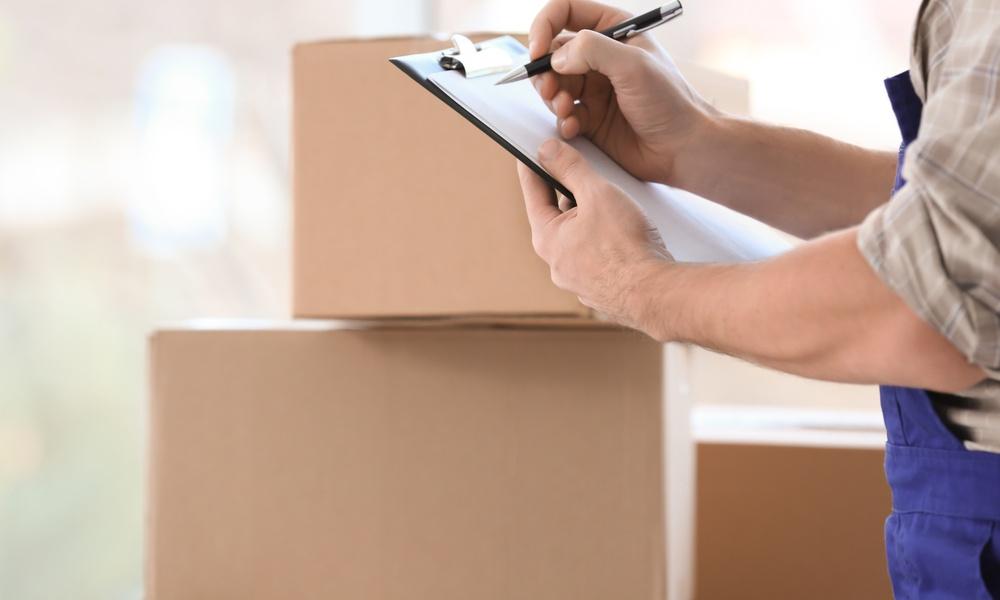 A man in blue overalls is writing on a piece of paper on a clipboard. There is a stack of cardboard boxes next to him.