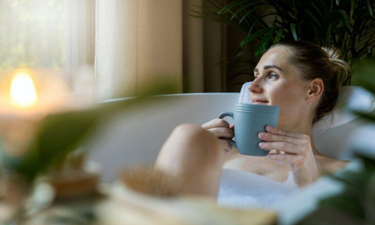 A blonde woman lying in a bathtub with a blue coffee mug in her hands. Indoor plants and a lit candle surround her.