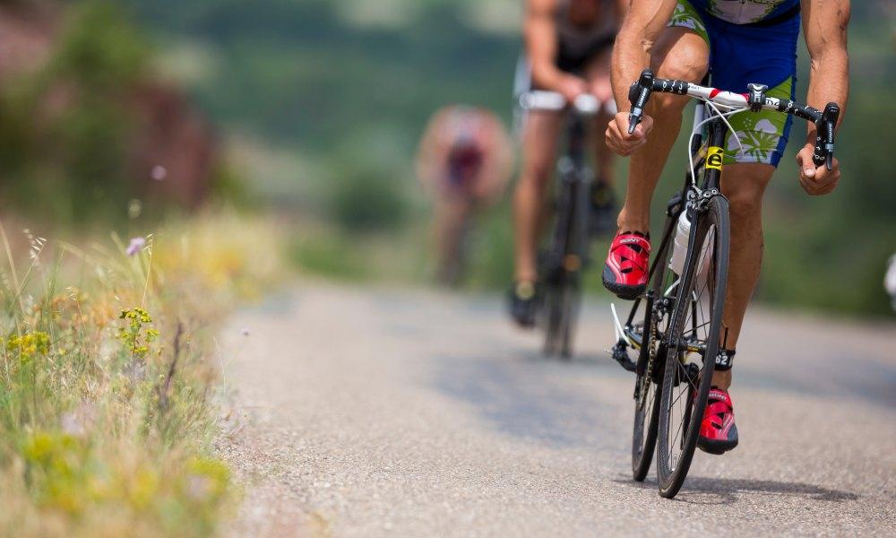 A ground-level view of competitive cyclists pedaling toward the viewer on a narrow concrete path surrounded by grass.
