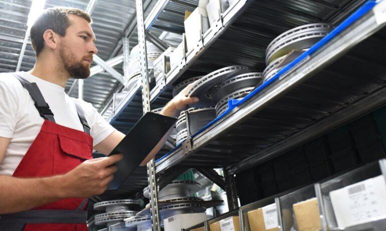 An employee in an auto parts store holding a tablet and removing a car part from a large metal shelving unit.