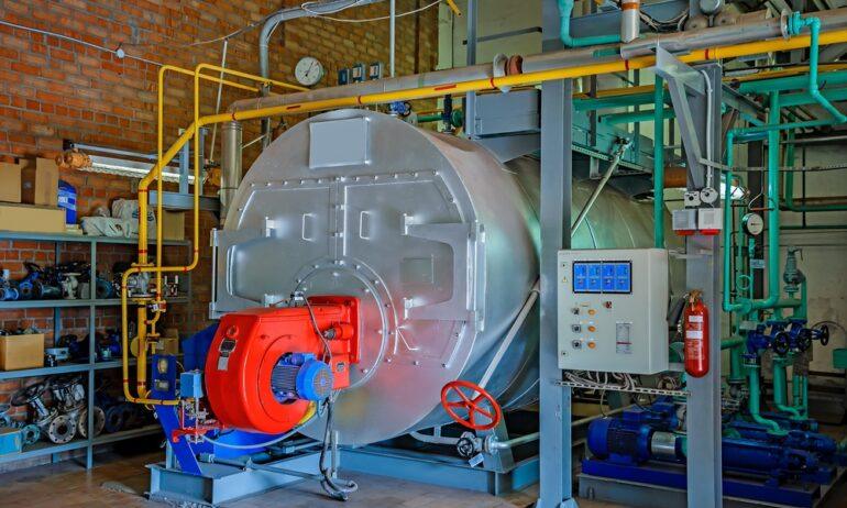 An industrial steam boiler room with exposed brick walls and a large stainless steel tank. The pipes are red and yellow.