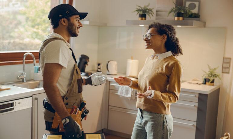 An electrician wearing gloves is smiling and talking to a homeowner in a kitchen. The homeowner is wearing glasses.