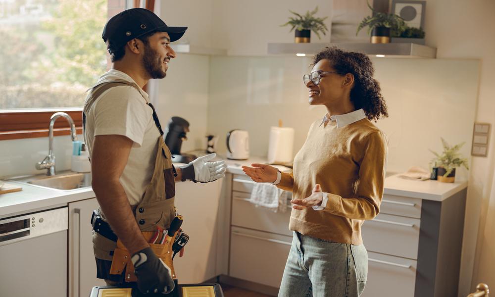 An electrician wearing gloves is smiling and talking to a homeowner in a kitchen. The homeowner is wearing glasses.