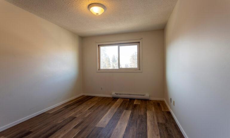 An empty bedroom with cream colored walls and vinyl plank flooring. There's a baseboard heater under the small window.