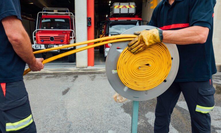 Two firefighters wrapping up a fire hose using a hose-wrapping machine. The garages behind them are both open.
