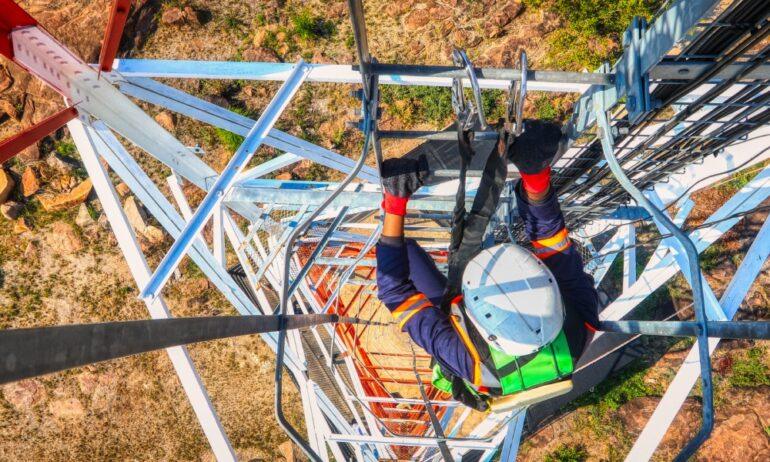 An aerial view of a worker climbing a steel communication tower. They are wearing a harness, hardhat, and a safety vest.